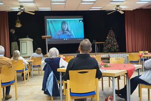 Ein paar Menschen sitzen an Tischen und schauen auf eine Leinwand auf der Bühne.