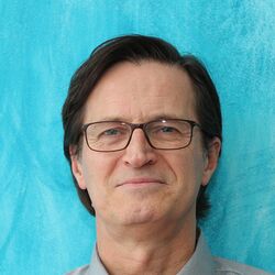 Eine Person mit kurzen schwarzen Haaren und Brille ist von vorne vor einem türkisfarbenen Hintergrund zu sehen.