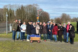 Einige Menschen stehen in Winterbekleidung mit einem Bollerwagen neben einer Wiese und Bahnschienen.