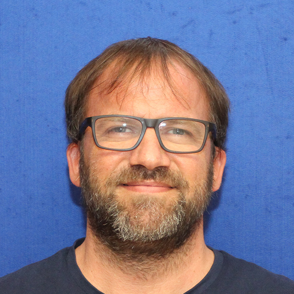 Eine Person mit brünetten Haaren und Brille ist vor einem blauen Hintergrund zu sehen.